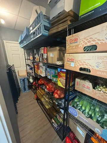 A stockroom aisle of wire shelving holding cereal boxes, tinned and boxed goods, tomatoes and crates of cucumbers in Dole banana boxes
